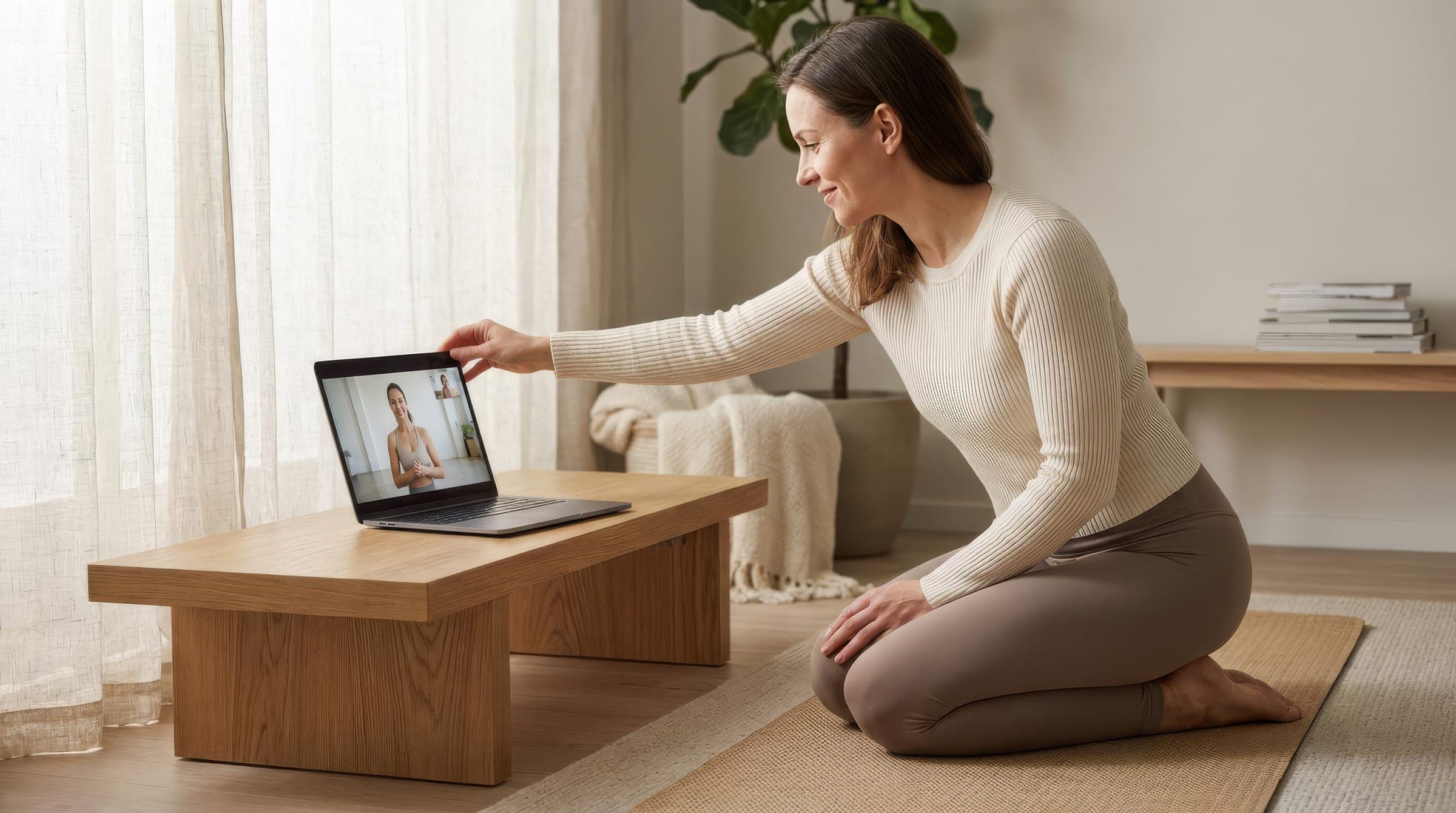 Frau kniet auf der Yogamatte vor einem Laptop und begrüßt ihre Trainerin im Video-Call